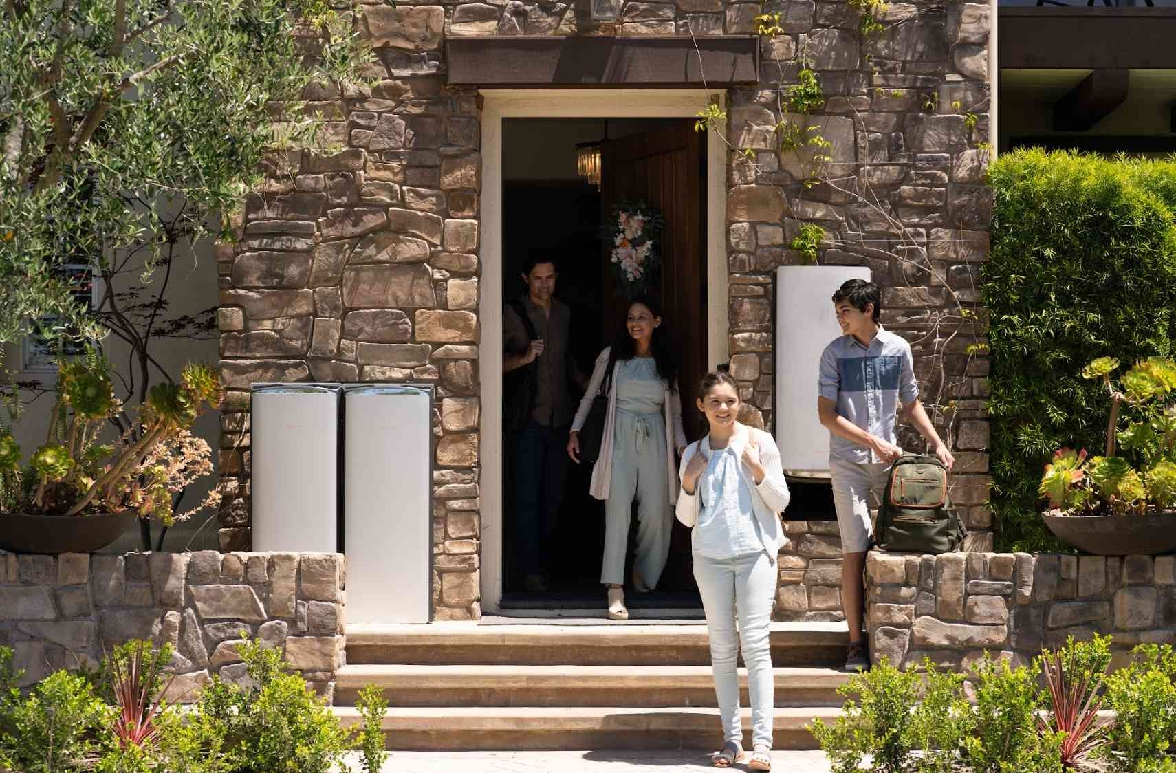 A family of four leaving their stone house through the front door, with two EcoFlow OCEAN Pro battery units visibly installed on the exterior wall next to the entrance.